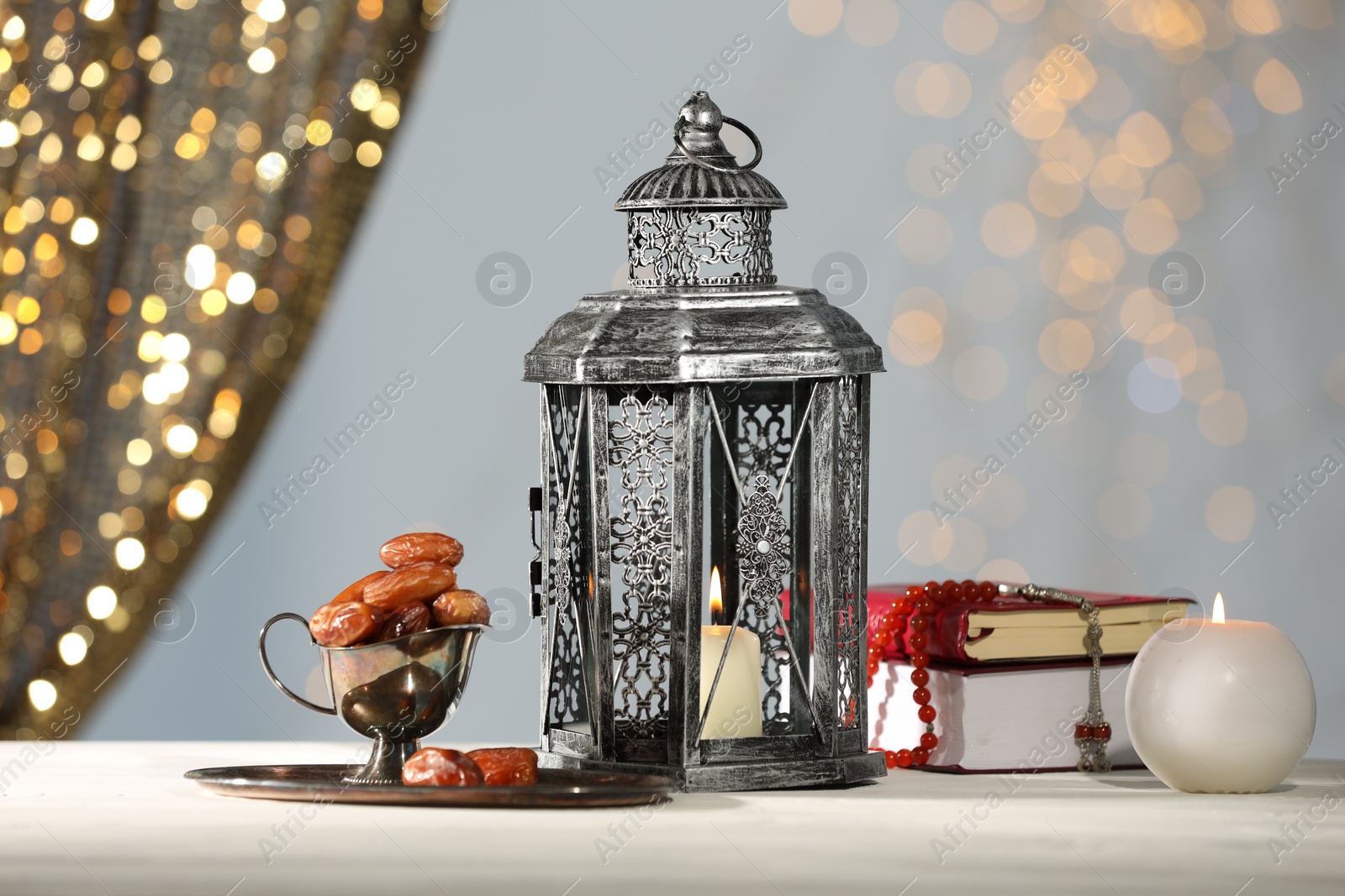 Photo of Arabic lantern, Quran, misbaha, candle and dates on white table against blurred lights