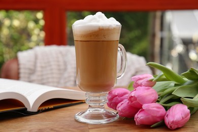 Glass of delicious cocoa, pink tulips and book on wooden table at terrace