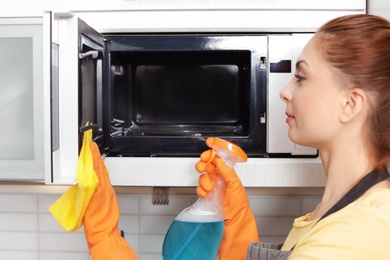 Woman cleaning microwave oven with rag and detergent in kitchen