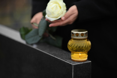 Photo of Woman holding white rose near black granite tombstone with candle outdoors, closeup. Funeral ceremony