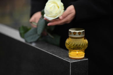 Woman holding white rose near black granite tombstone with candle outdoors, closeup. Funeral ceremony