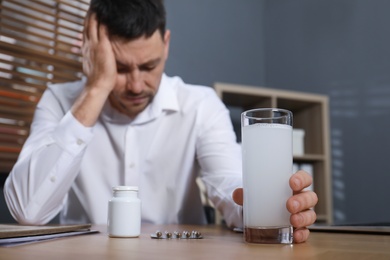 Man taking medicine for hangover at desk in office, focus on hand with glass