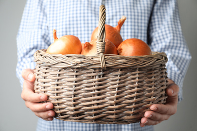 Photo of Woman holding wicker basket with raw yellow onion bulbs on grey background, closeup