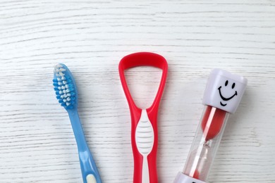 Photo of Tongue cleaner, toothbrush and hourglass on white wooden table, flat lay