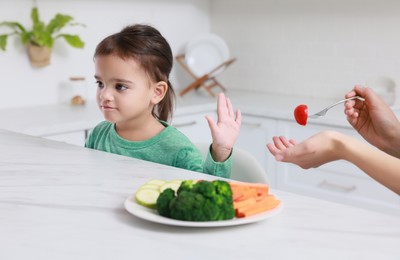 Mother feeding her daughter in kitchen, closeup. Little girl refusing to eat vegetables