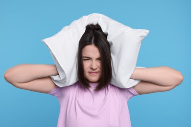 Photo of Irritated young woman covering ears with pillow on light blue background. Insomnia problem