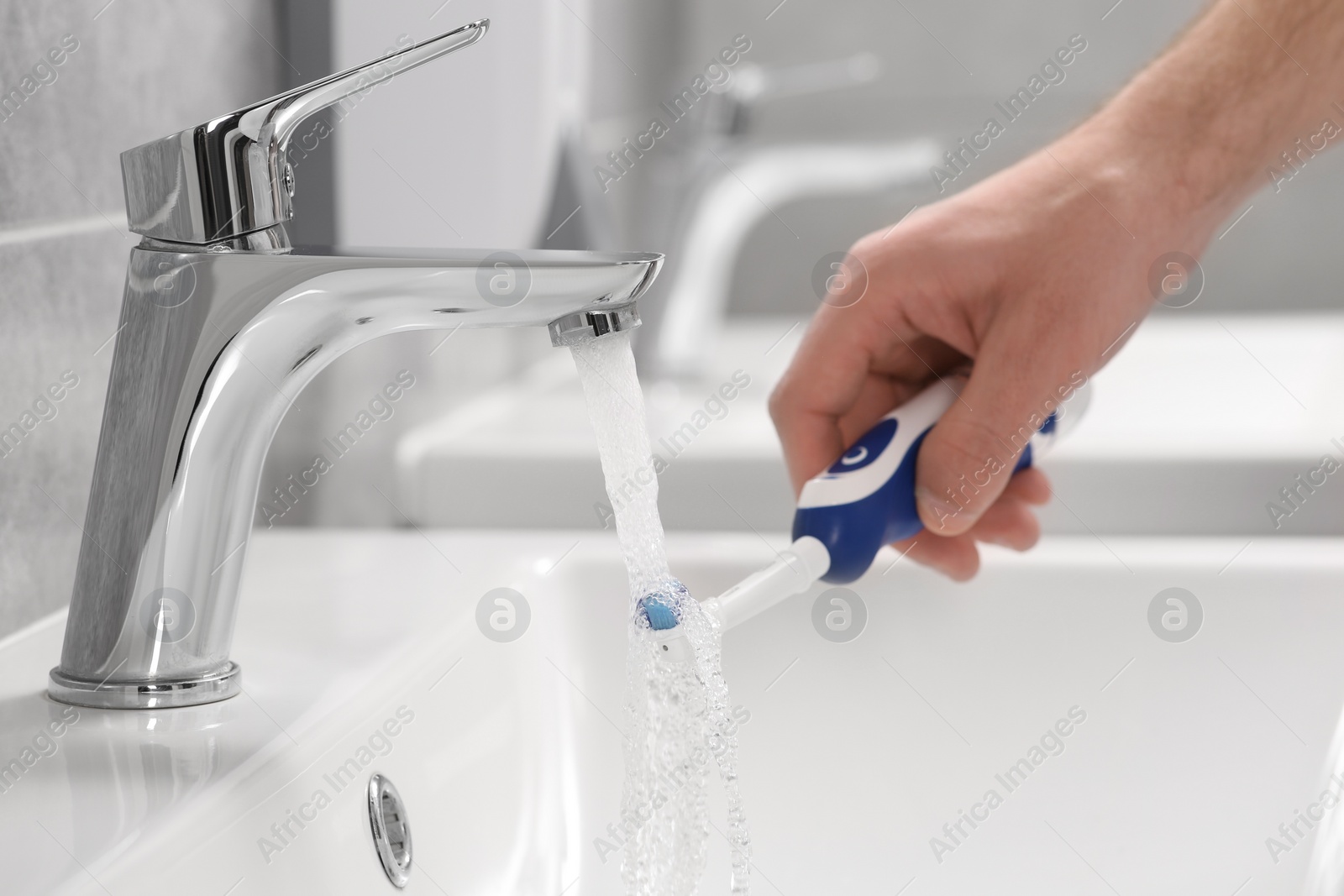 Photo of Man holding electric toothbrush under flowing water above sink in bathroom, closeup