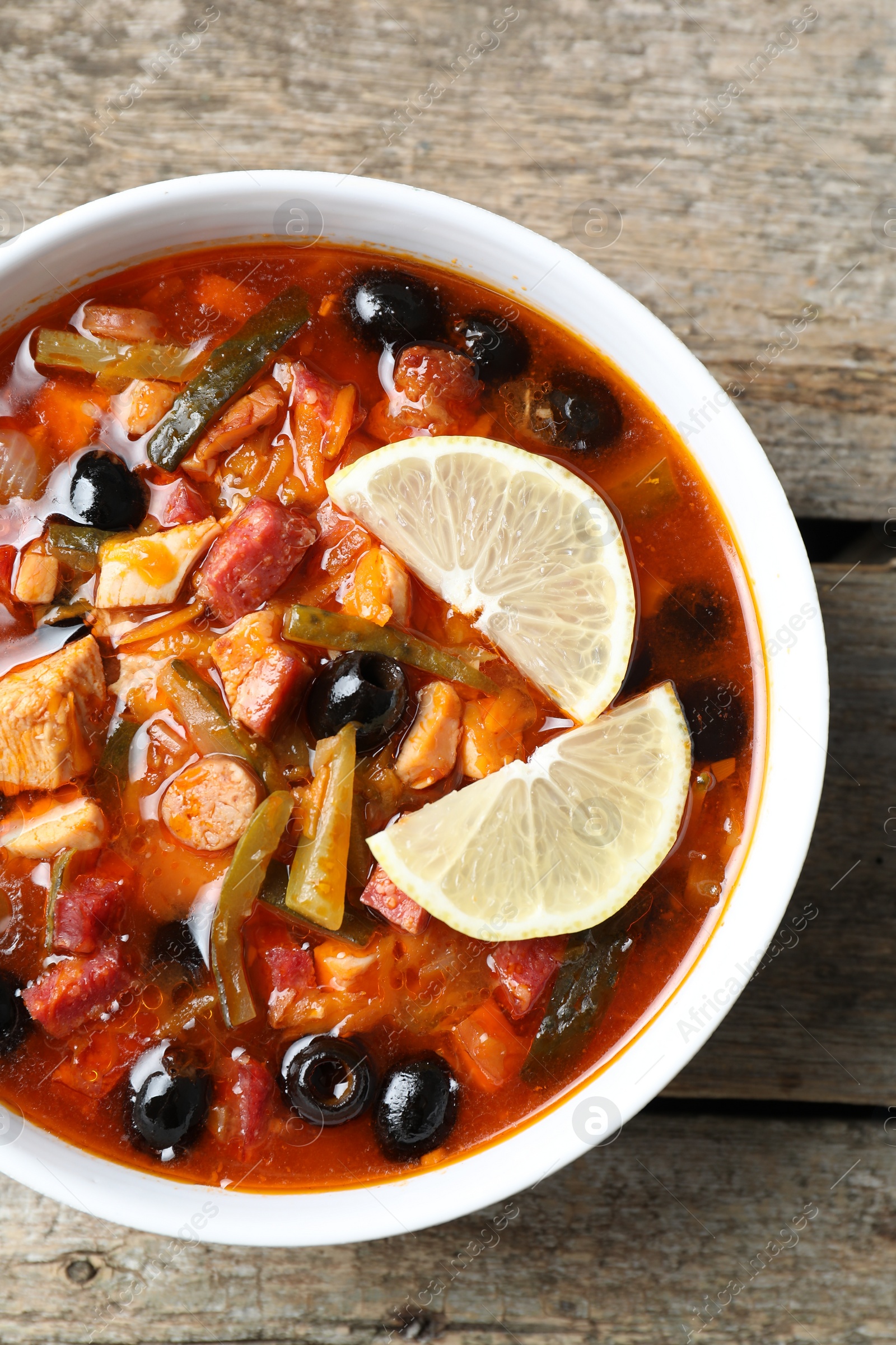 Photo of Meat solyanka soup with sausages, olives and vegetables in bowl on wooden table, top view