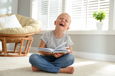 Photo of Cute little boy reading book on floor at home