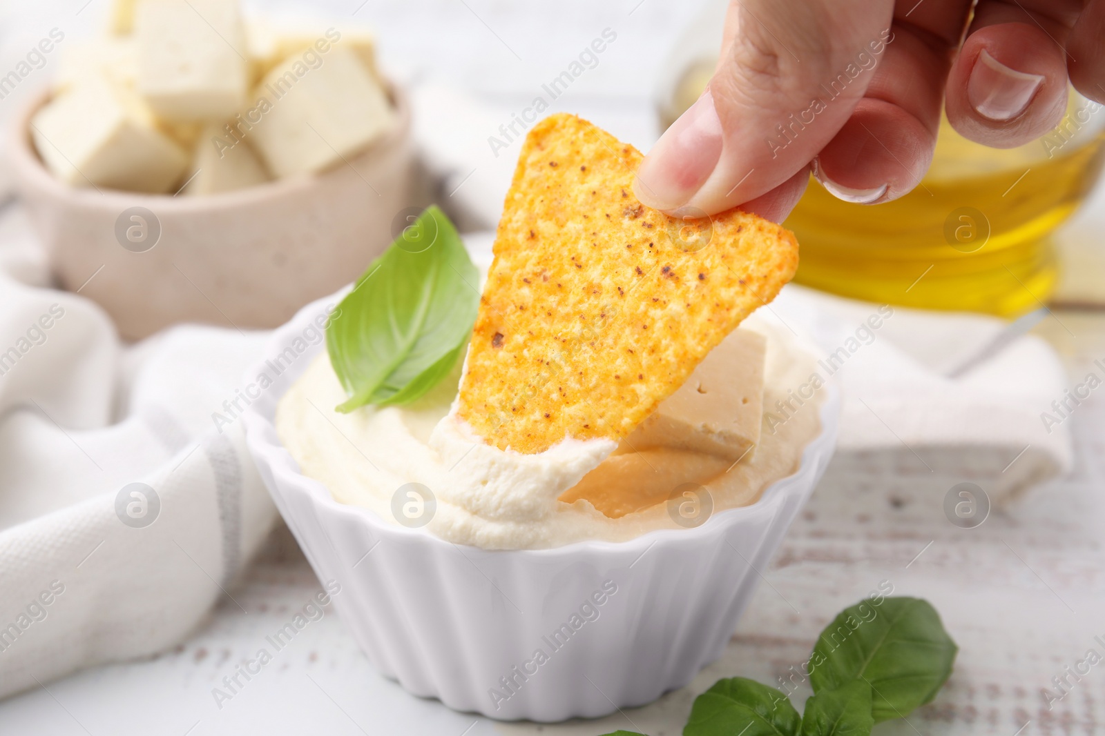 Photo of Woman dipping nachos chip into delicious tofu sauce at white wooden table, closeup