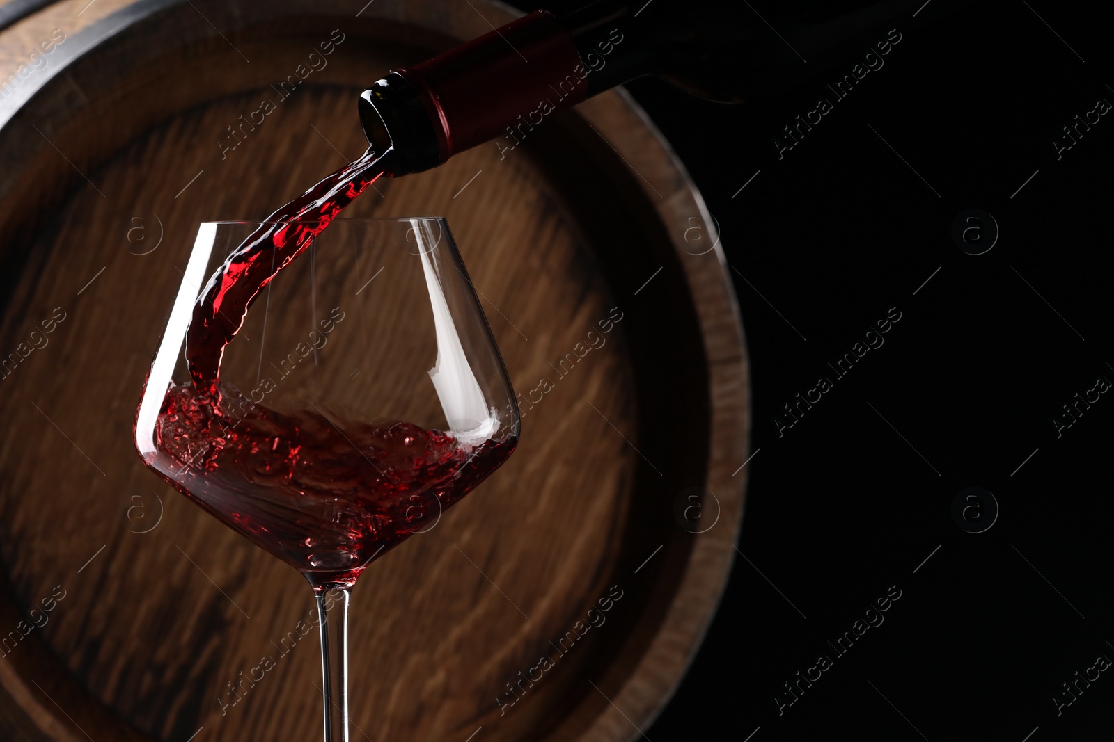 Photo of Pouring red wine from bottle into glass near wooden barrel against black background. Space for text