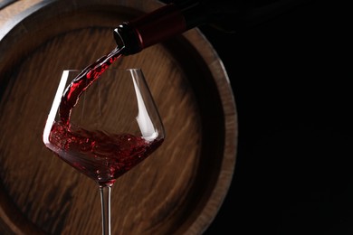 Photo of Pouring red wine from bottle into glass near wooden barrel against black background. Space for text