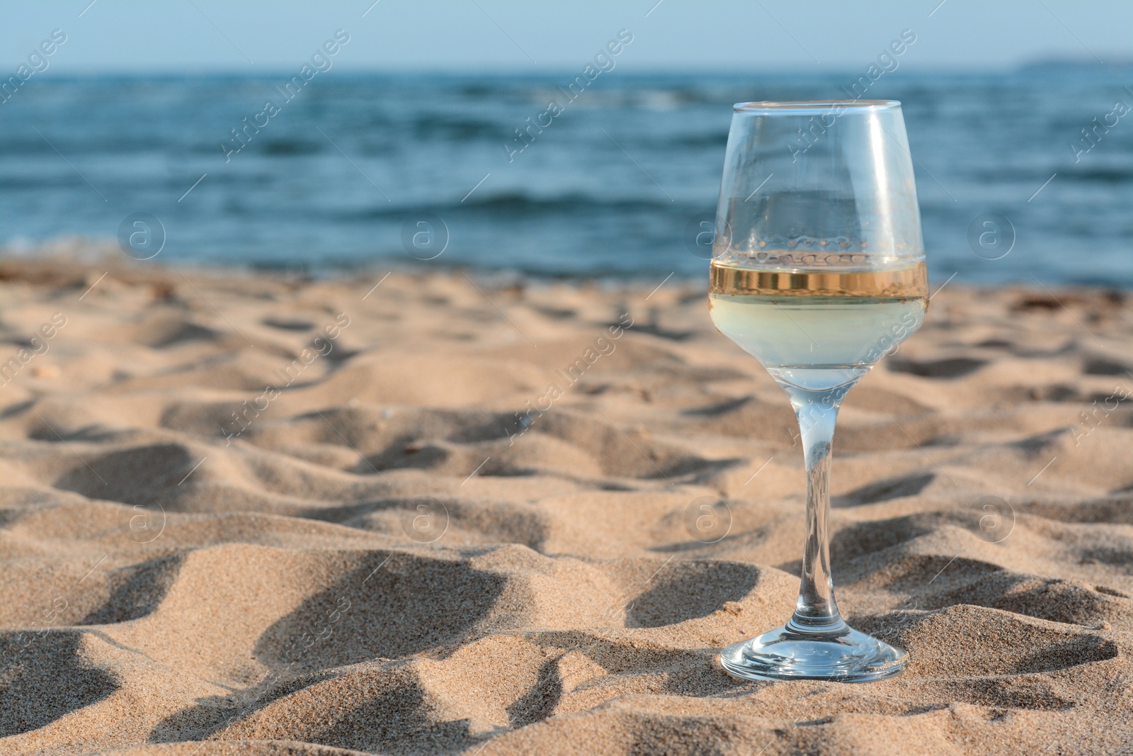 Photo of Glass of tasty wine on sand near sea, space for text