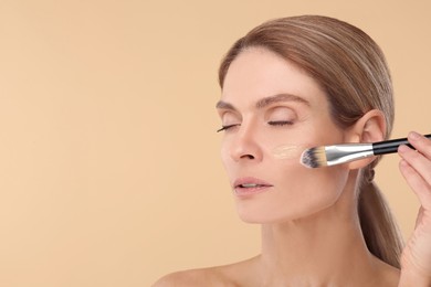 Woman applying foundation on face with brush against beige background. Space for text