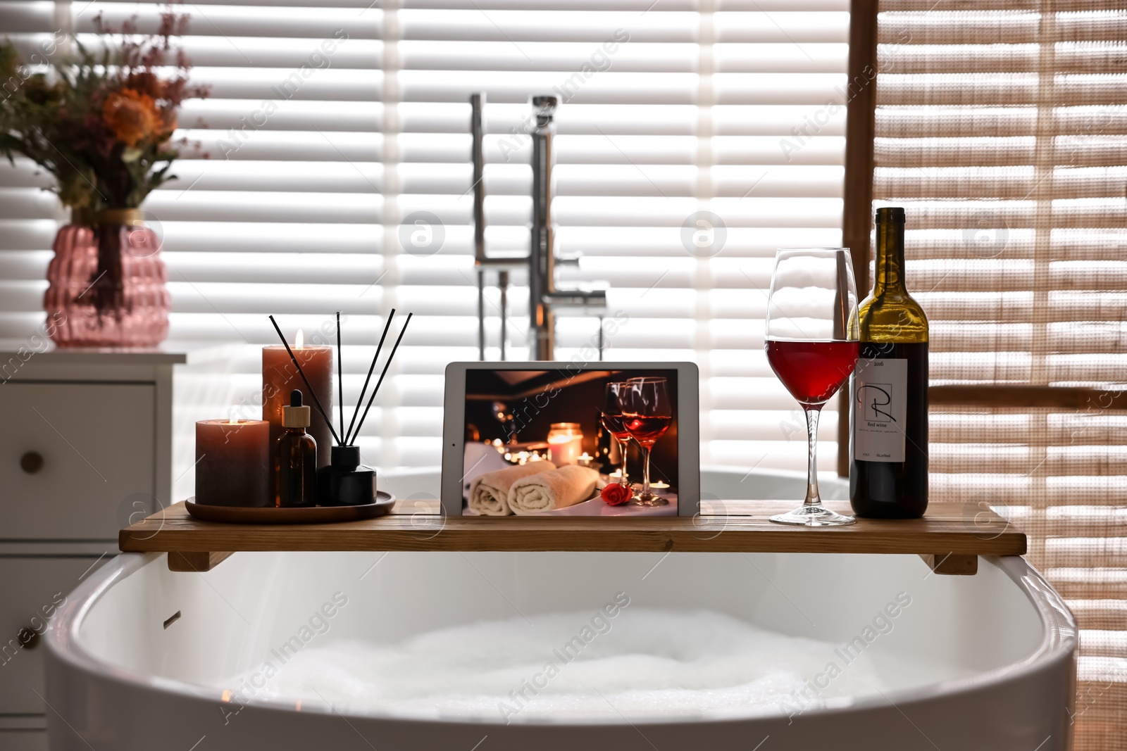 Photo of Wooden tray with tablet, wine and burning candles on bathtub in bathroom