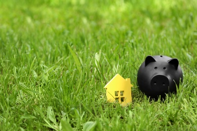 Photo of Black piggy bank and model of house in green grass outdoors. Space for text