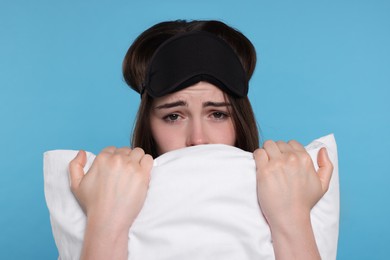 Unhappy young woman with sleep mask and pillow on light blue background. Insomnia problem