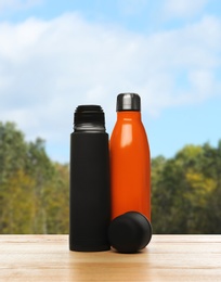 Thermos bottles on wooden table against blurred forest