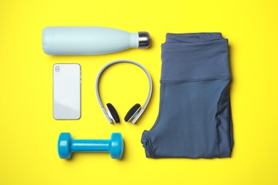 Photo of Stylish sports leggings, dumbbell, thermo bottle, headphones and smartphone on yellow background, flat lay