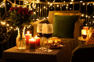 Photo of Vase with roses, glasses of wine, snacks and candles on rattan table at balcony in night