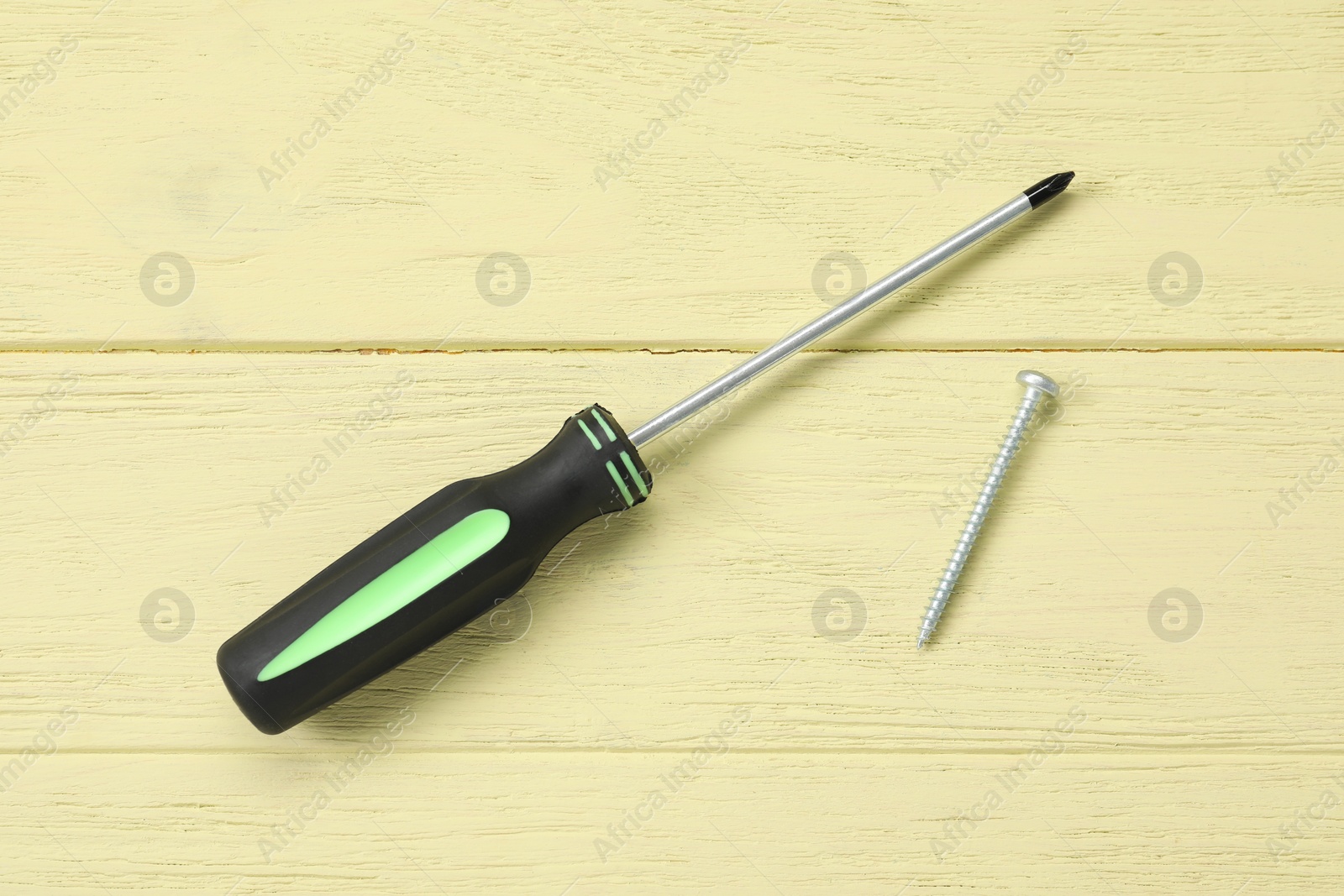 Photo of Screwdriver with black handle and screw on yellow wooden table, flat lay