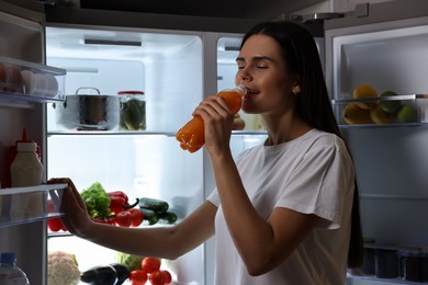 Photo of Young woman drinking juice near modern refrigerator at night