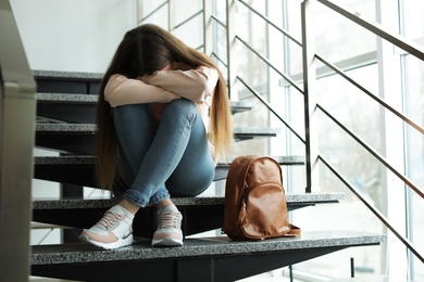 Photo of Upset teenage girl with backpack sitting on stairs indoors
