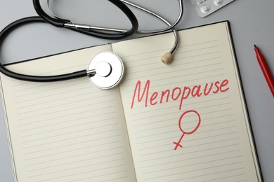 Photo of Stethoscope near notebook with word Menopause and female gender sign on light grey background, flat lay