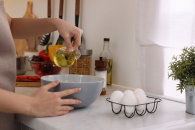 Photo of Cooking process. Woman pouring oil from bottle into bowl at light countertop indoors, closeup