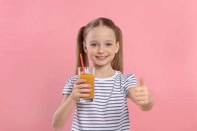 Photo of Cute little girl holding glass of fresh juice with straw and showing thumbs up on pink background