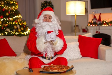 Photo of Merry Christmas. Santa Claus with cupdrink watching TV on sofa at home