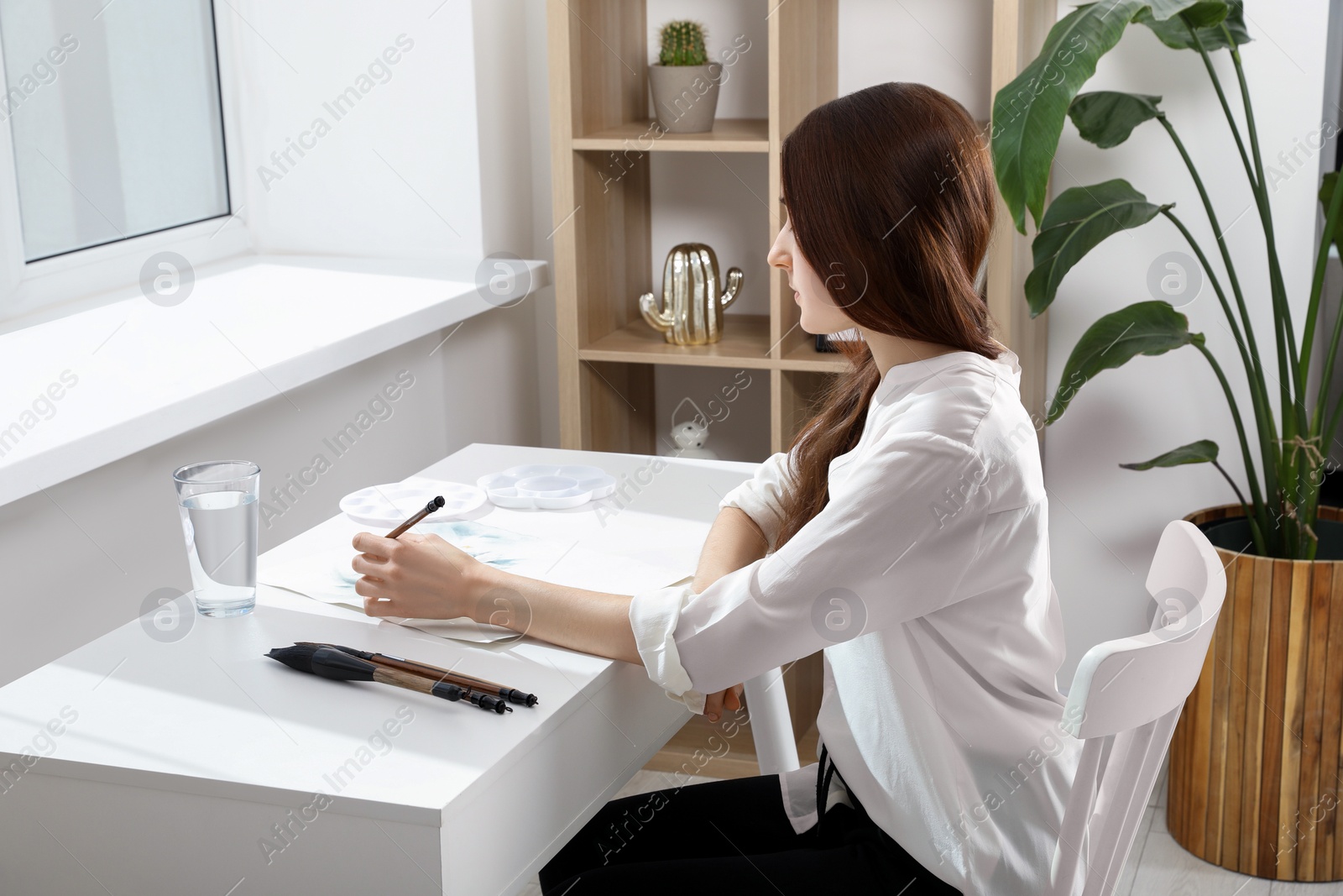 Photo of Woman painting flowers with watercolor at white table in room. Creative artwork