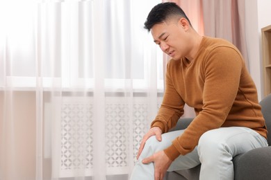 Photo of Asian man suffering from knee pain on sofa indoors