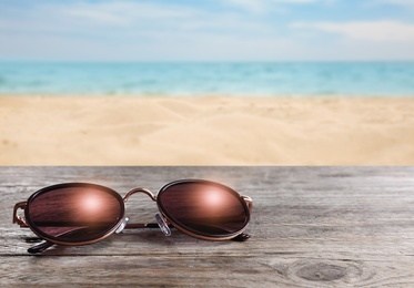 Image of Stylish sunglasses on wooden table near sea with sandy beach