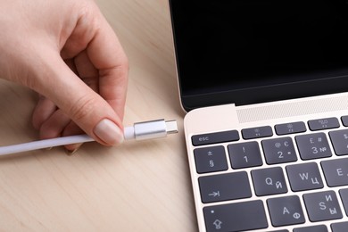 Photo of Woman plugging USB cable with type C connector into laptop port on light wooden table, closeup