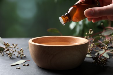 Photo of Woman dripping eucalyptus essential oil from bottle into bowl at grey table, closeup