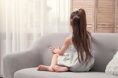 Photo of Lonely little girl sitting on sofa near window indoors. Child autism