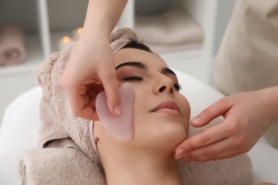 Photo of Young woman receiving facial massage with gua sha tool in beauty salon