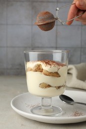 Photo of Woman dusting delicious tiramisu with cocoa powder on light table, closeup