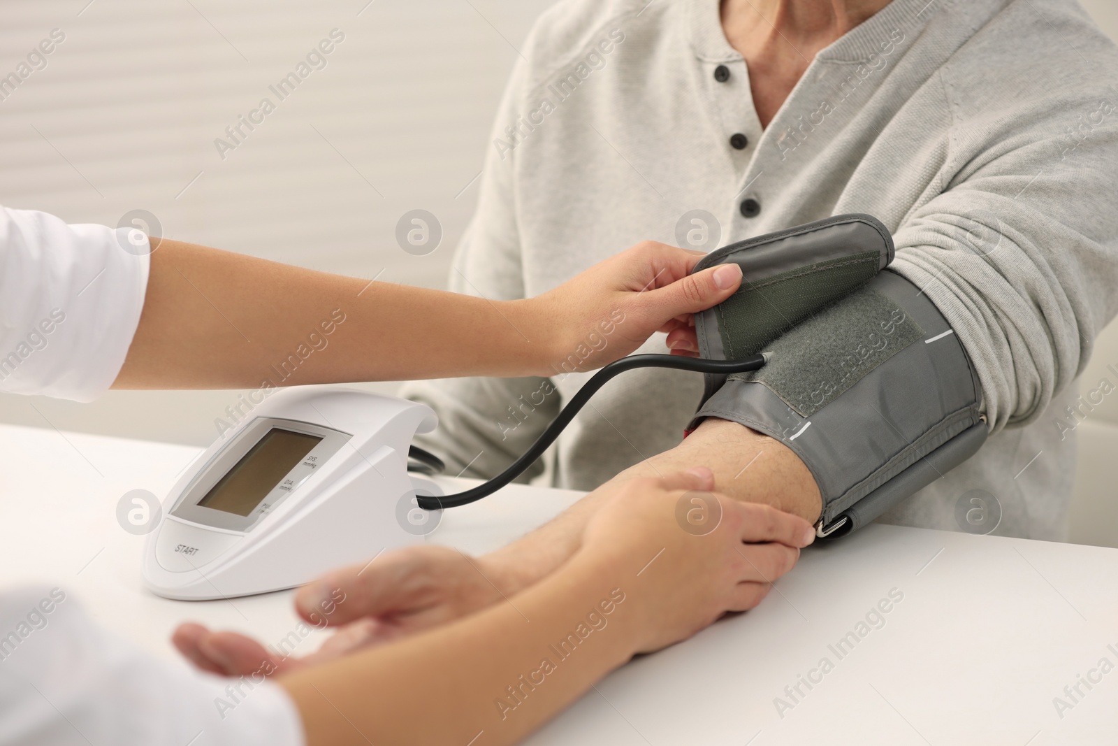 Photo of Nurse measuring elderly patient's blood pressure at white table indoors, closeup