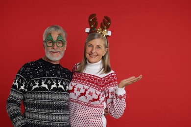 Photo of Senior couple in Christmas sweaters, reindeer headband and party glasses on red background. Space for text