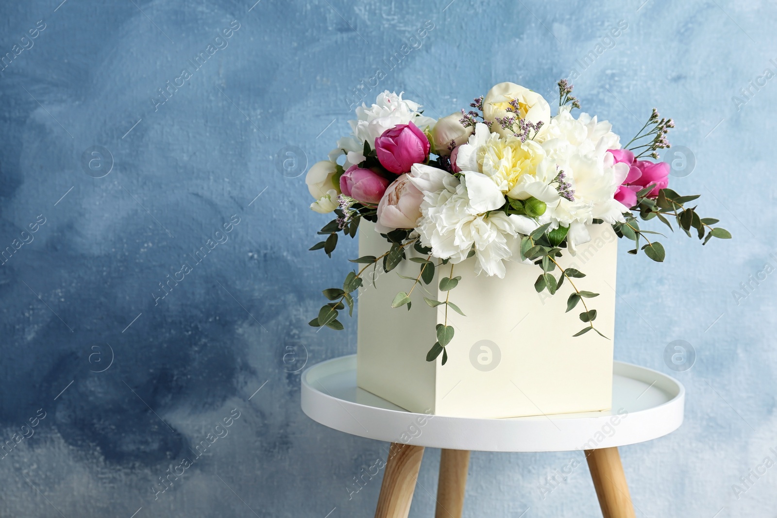 Photo of Box with bouquet of beautiful flowers on table against color background