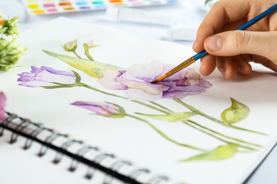 Photo of Woman painting eustomas in sketchbook at white table, closeup