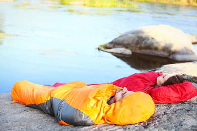 Young couple resting in sleeping bags on cliff near lake