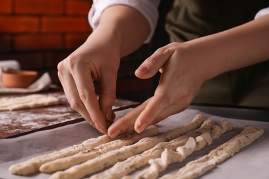 Photo of Woman putting homemade breadsticks on baking sheet indoors, closeup. Cooking traditional grissini