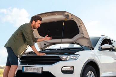 Photo of Upset man looking under hood of broken car outdoors, low angle view