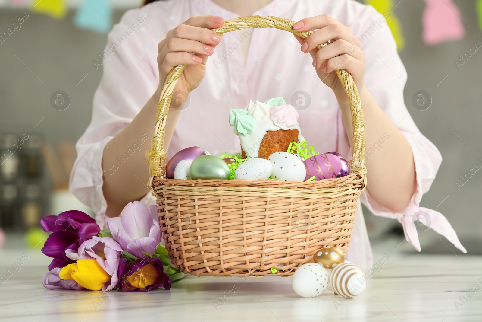 Photo of Woman holding wicker basket with painted eggs and delicious Easter cake near tulips at white marble table indoors, closeup