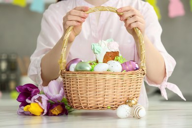 Woman holding wicker basket with painted eggs and delicious Easter cake near tulips at white marble table indoors, closeup