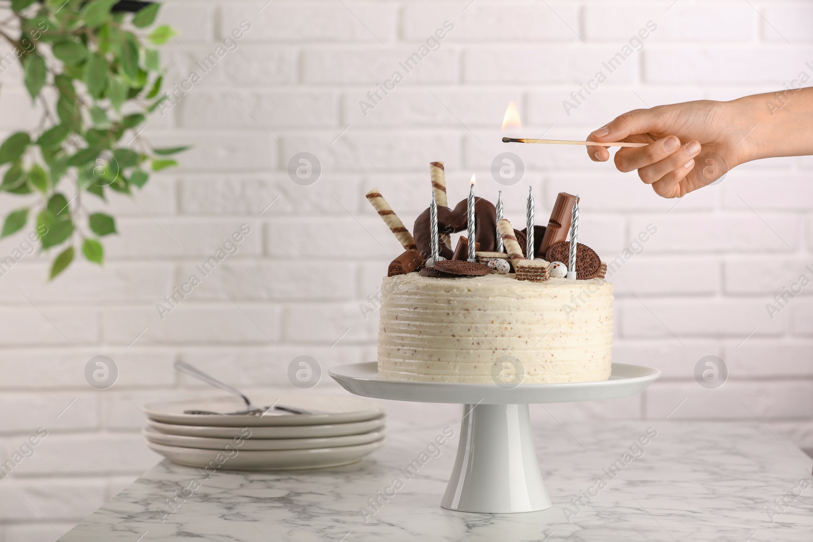 Photo of Woman lighting candle on delicious cake decorated with sweets at white marble table, closeup. Space for text