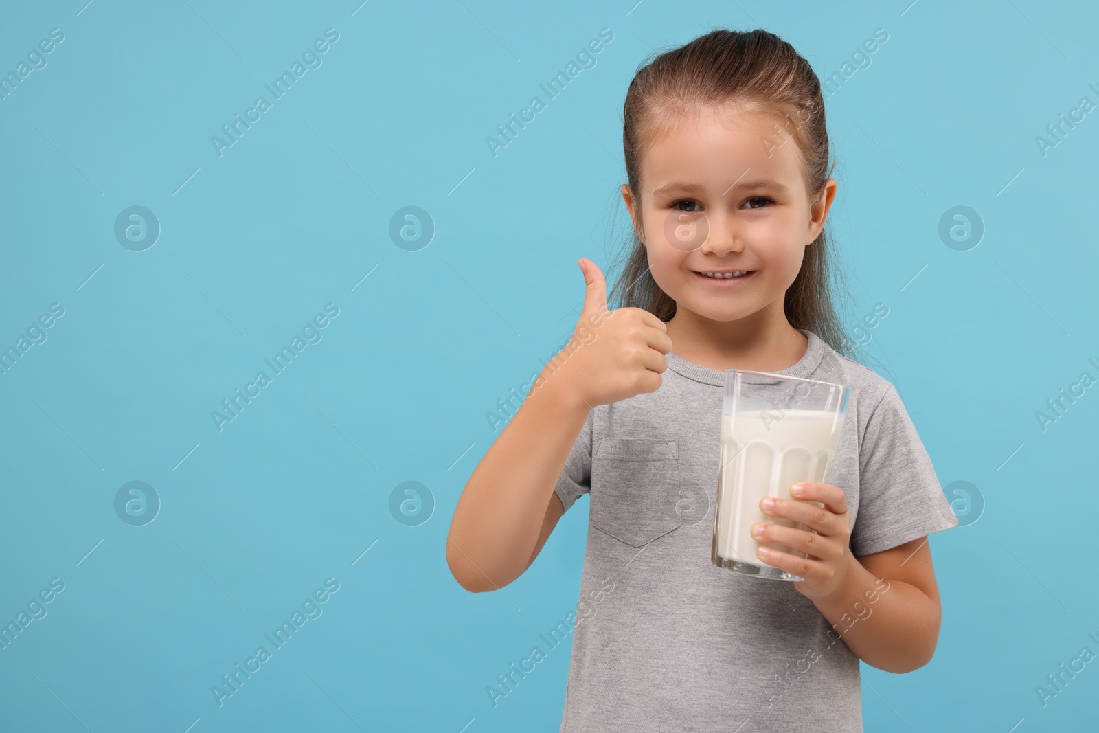 Photo of Cute girl with glass of fresh milk showing thumb up on light blue background, space for text
